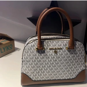 Michael Kors handväska med MK-mönster - Snygg handväska från Michael Kors i beige och brunt med klassiskt MK-mönster över hela väskan. Väskan har två rejäla handtag, guldfärgade detaljer och flera fack med dragkedja. Tillverkad i skinn och syntetmaterial, perfekt för dig som gillar stilrena accessoarer.
