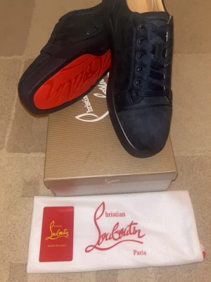 Svarta sneakers från Christian Louboutin - Snygga svarta sneakers från Christian Louboutin med klassisk röd sula och rund tå. Skorna är tillverkade i mocka och har snörning framtill. Perfekt för dig som vill ha en lyxig och stilren look med exklusiva detaljer.