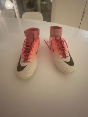 Nike Mercurial fotbollsskor rosa/vit - Säljer ett par Nike Mercurial fotbollsskor i en snygg rosa och vit färg med svarta detaljer och snörning. Skorna har hög strumpkrage och platt sula med dobbar för gräsplan. Materialet är syntet och skon har en rund tå. Perfekt för dig som vill sticka ut på planen!