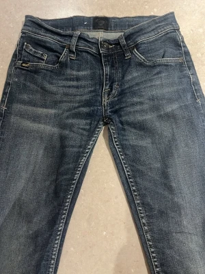 Mörkblå jeans från Tiger of Sweden - Snygga mörkblå jeans från Tiger of Sweden med klassisk femficksdesign och kontrastsömmar. Jeansen har slim passform och raka ben, med detaljer som knappstängning och läderpatch med logga bak i midjan. Perfekta för dig som gillar stilrena och tidlösa jeans.