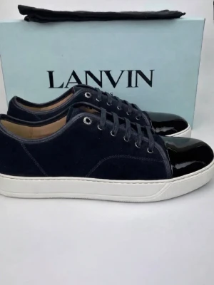 Mörkblå sneakers från Lanvin - Snygga mörkblå sneakers från Lanvin med vit platt sula och glansig svart tå. Skorna har snörning och är tillverkade i mocka med detaljer i lack. Perfekta för dig som gillar stilrena och exklusiva sneakers. Köptes för en månad sen från care of Carl allt ingår!! Kan sänka priset vid snabb affär!!