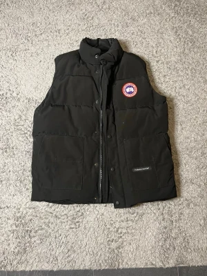Svart dunväst från Canada Goose - Svart dunväst från Canada Goose med hög krage, två stora fickor framtill och klassisk logotyp på bröstet. Västen är o storlek M och är i mycket bra skick, används dock ej längre. Pris kan diskuteras vid snabb affär 😊