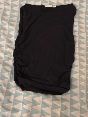 Svart topp med rynkade sidor - Säljer en svart topp med snygga rynkade detaljer på ena sidan. Toppen är axelbandslös och har en enkel, stilren design som är lätt att matcha. Materialet känns mjukt och stretchigt, perfekt för dig som gillar minimalistisk stil.