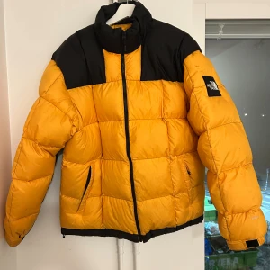 Gul och svart pufferjacka North Face - Säljer en ikonisk gul och svart pufferjacka från The North Face. Jackan har hög krage, dragkedja framtill och två fickor med dragkedja. Klassisk boxig passform och stora quiltade partier. Perfekt för kalla dagar och riktigt snygg streetstil.