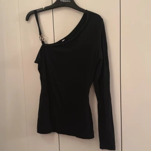 Svart oneshoulder topp från Bubbleroom - Snygg svart oneshoulder topp från Bubbleroom med lång ärm och en axelbandslös sida. Toppen har ett smalt axelband med guldfärgad metallring med CF-logga(ciara forthi gissar jag). Materialet är mjukt och stretchigt, perfekt för att sticka ut lite extra. Strl står ej men den passar XS/S skulle jag säga. 