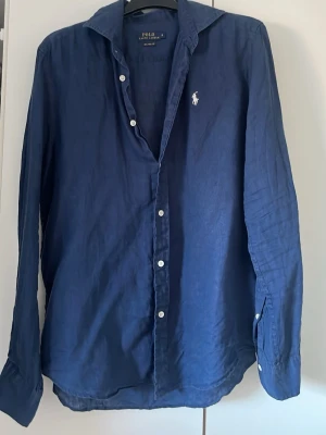 Blå linneskjorta från Polo Ralph Lauren - Snygg marinblå skjorta från Polo Ralph Lauren i linne med klassisk krage och vita knappar. Långärmad modell med avslappnad passform och ikonisk broderad logga på bröstet. Perfekt för dig som gillar stilrena och bekväma plagg. 