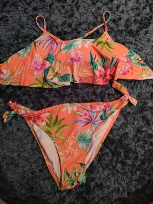 Blommigt bikini set från Calzedonia - Säljer en färgglad bikini från Calzedonia Baby Sea i storlek 12 år. Bikinin har ett tropiskt blommigt mönster i orange, rosa, grönt och lila med volang upptill och knyt i sidorna på underdelen. Materialet är mjukt och stretchigt syntet, perfekt för strandhäng.