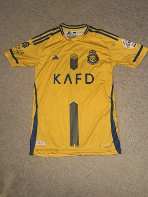 Al nassr tröja - Bilderna visar en gul fotbollströja från Al Nassr med blå detaljer och tryck, inklusive namn och nummer på ryggen. Ingen sko finns med på bilderna.