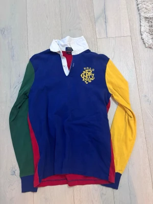 Färgglad långärmad pikétröja Polo Ralph Lauren - Säljer en färgstark långärmad pikétröja från Polo Ralph Lauren med broderad logga på bröstet. Tröjan har blå front, röd rygg, grön och gul ärm, vit krage och blå muddar. Tillverkad i mjuk bomull, perfekt för dig som gillar sportig och retro stil.