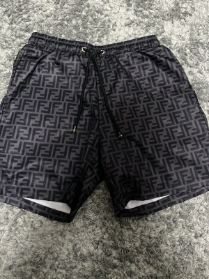 Svarta Fendi badshorts med snörning - Säljer ett par svarta Fendi badshorts med all-over logomönster och snörning i midjan. De har en loose passform och är gjorda i ett syntetmaterial som känns lätt och skönt. Perfekta för dig som gillar designerplagg med tydlig branding.