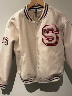 Beige bomberjacka - Snygg beige bomberjacka från H&M med stor röd S-applikation och siffran 96 på ärmen. Jackan har ribbade muddar med rödblåa ränder, tryckknappar framtill och två sidofickor. 