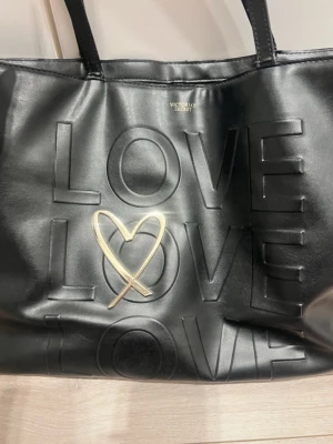 Svart shoppingväska från Victoria's Secret - Säljer en svart shoppingväska från Victoria's Secret med präglad text 'LOVE' och ett guldfärgat hjärta på framsidan. Väskan har två rejäla handtag och är tillverkad i skinnimitation. Perfekt för att bära med sig allt man behöver under dagen.