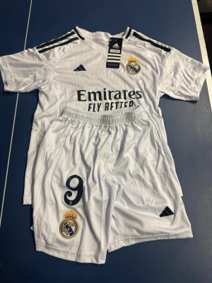 Real Madrid fotbollströja Mbappé #9 - 9-10 år - Säljer en vit Real Madrid fotbollströja med Mbappé och nummer 9 på ryggen. Setet inkluderar matchande shorts med klubbens emblem och Adidas-logga. Tröjan har svarta detaljer och klubbmärke på bröstet. Perfekt för dig som älskar fotboll och Real Madrid. 9-10 år storlek