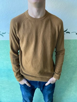 Brun stickad tröja från Riley - Säljer en brun stickad tröja från Riley i storlek M. Tröjan har ett diskret strukturmönster, rund hals och ribbade muddar vid ärmslut och nederkant. Perfekt för dig som gillar enkel och stilren design med lite extra textur.