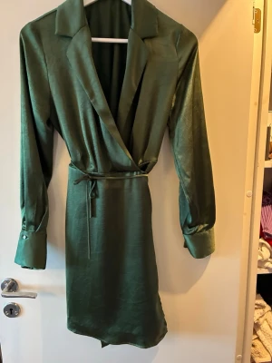 Grön ’satin’ omlottklänning från Mango  - Snygg grön omlottklänning från MNG i satinliknande material. Klänningen har långa ärmar med glittriga knappar vid manschetterna, klassisk krage och ett smalt bälte i midjan. Perfekt för dig som gillar stilrena detaljer och en elegant look.
