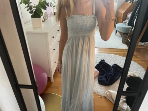 Ljusblå långklänning med smock - Superfin långklänning i ljusblått och vitt med smala ränder. Klänningen har smock upptill, tunna axelband och volangkant vid bröstet. Materialet känns lätt och luftigt, perfekt för varma dagar. Passformen är normal och klänningen är ärmlös. Så himla fin! Som ny! 