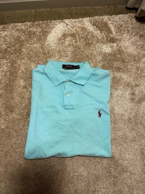 Turkos Polo Ralph Lauren piké - Snygg turkos pikétröja från Polo Ralph Lauren i custom slim fit. Klassisk krage, två knappar och ikonisk röd broderad logga på bröstet. Vinröd logo! Tillverkad i mjuk bomull som är skön mot huden. Perfekt för dig som gillar stilrena och fräscha plagg. Priset ej huggget i sten hör av er vid frågor eller funderingar! 