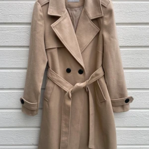 Beige kappa från Fisherfield med bälte - Stilren beige kappa från Fisherfield med klassisk krage, knytskärp i midjan och svarta knappar framtill. Jackan är helfodrad och har långa ärmar med knappdetalj vid ärmslut. Så snygg och knappt använd pga för liten Obs! Mer lik stl. 36 än 38