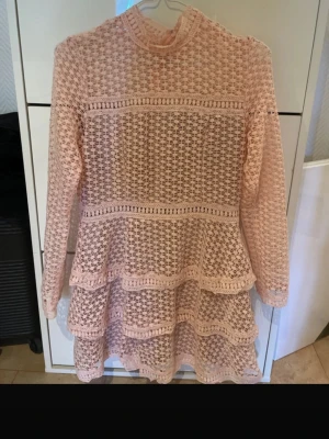 Ljusrosa spetsklänning från AX Paris - Superfin ljusrosa spetsklänning från AX Paris med lång ärm och volangdetaljer. Klänningen har ett genombrutet spetsmönster, hög hals och dragkedja i sidan. Perfekt för dig som vill ha en söt och romantisk look.