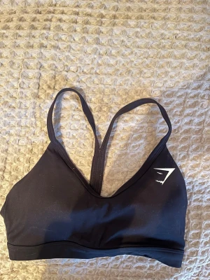 Svart sport-bh från Gymshark - Svart sport-bh från Gymshark i storlek XS med smala axelband och snyggt racerback-rygg. Materialet är mjukt och stretchigt, perfekt för träning. Diskret logga i vitt framtill. Sitter bekvämt och ger bra stöd under intensiva pass. Endast använd någon enstaka gång.