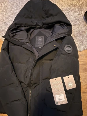 Svart pufferjacka från Canada Goose - Säljer en svart pufferjacka från Canada Goose med huva och diskret logotyp på ärmen. Jackan har dragkedja och tryckknappar framtill samt flera praktiska innerfickor. Perfekt för kalla dagar och riktigt snygg. Den är i storlek M men passar S lika bra. Den har QR kod skanning direkt till hemsidan med NFC scanning också.