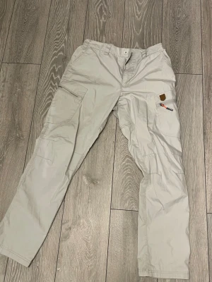 Beige cargopants från Tuxer - Säljer ett par beige cargopants från Tuxer med flera praktiska fickor och coola detaljer som orange dragkedja. Byxorna har rak passform och är gjorda i slitstarkt syntetmaterial, perfekta för dig som gillar funktion och stil.