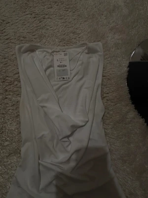 Vit omlott ärmlös topp från Stradivarius - Säljer en snygg vit omlottopp från zara i storlek S. Toppen har djup v-ringning och är ärmlös med ett draperat fall framtill. Materialet är mjukt och stretchigt, perfekt för att skapa en clean och fräsch look. Prislappen sitter kvar.