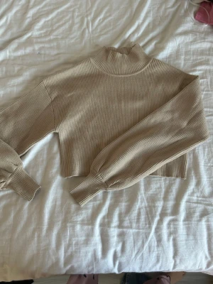 Beige croppad stickad polotröja Gina Tricot - Säljer en beige croppad stickad polotröja från Gina Tricot i storlek XS. Tröjan har ribbad struktur, lång ärm med bred mudd och hög krage. Perfekt för dig som gillar en enkel men trendig look med lite loose passform.