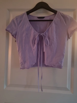 Ljuslila croppad topp med knyt - Säljer en ljuslila croppad topp med korta ärmar och v-ringning. Toppen har en snygg knytning framtill som ger en trendig look. Materialet känns mjukt och är troligtvis bomull eller en bomullsblandning. Perfekt att styla med jeans eller kjol för en chill vibe.
