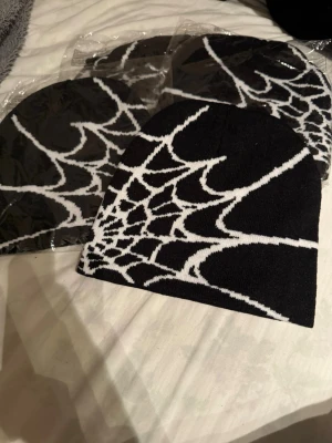 Svart mössa med spindelnätsmönster - Cool svart mössa med vitt spindelnätsmönster som täcker hela ytan. Stickad i mjukt material som sitter skönt på huvudet och ger en edgy vibe. Perfekt för dig som gillar unika accessoarer och vill sticka ut lite extra. Helt nya finns flera