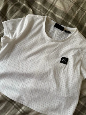 Vit mesh topp från Calvin Klein - Säljer en vit mesh topp från Calvin Klein Performance. Toppen har korta ärmar, rund hals och ett croppat snitt. Framsidan har en liten svart CK-logga och baksidan har ett vitt band med CK-tryck längs ryggen. Perfekt för träning eller chill.