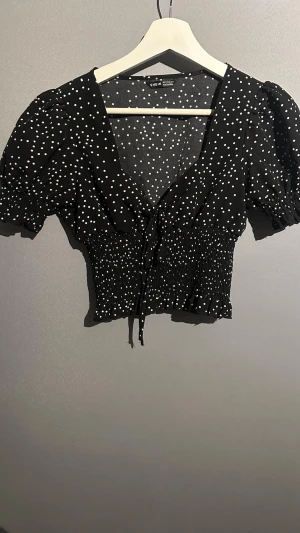 Svart prickig blus från SHEIN - Säljer en svart blus från SHEIN med vita prickar, v-ringad hals och kort puffärm. Toppen har smockad midja och knyt framtill för extra snygg passform. Perfekt till jeans eller kjol för en trendig look.
