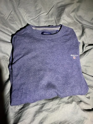 Marinblå stickad tröja från GANT - Säljer en marinblå stickad tröja från GANT i storlek S. Tröjan har rund halsringning, ribbade muddar och GANT-logga med sköld broderad på bröstet. Perfekt till lager på lager och enkel att matcha med jeans eller chinos.