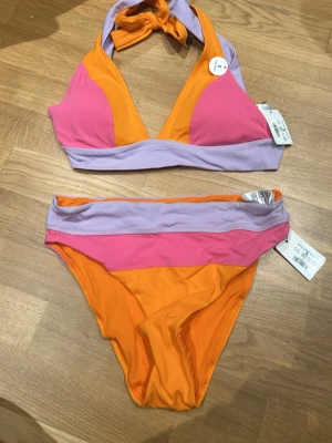 Färgglad bikini i orange, rosa och lila - Säljer en bikini i starka färger från Kappahl. Överdelen har trekantskupor med breda band i lila och knyt i nacken. Både över- och underdel har blockfärger i orange, rosa och lila. Materialet är mjukt och stretchigt, perfekt för strandhäng. Nypris 449kr