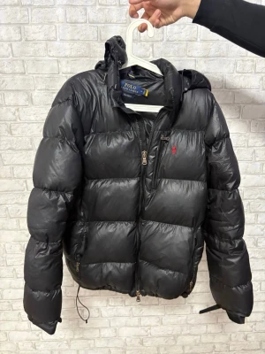 Svart pufferjacka från Polo Ralph Lauren - Säljer en svart pufferjacka från Polo Ralph Lauren med huva och dragkedja framtill. Jackan har en klassisk pufferdesign med breda kanaler, två sidofickor med dragkedja och Polo-logga broderad i rött på bröstet. Perfekt för kyliga dagar. Osäker på äkthet. Hör av dig vid intresse