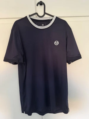 Mörkblå t-shirt från Sergio Tacchini - Säljer en mörkblå t-shirt från Sergio Tacchini med vit ribbad krage och diskret perforerat mönster över bröstet. T-shirten har korta ärmar och en snygg logga på framsidan. Perfekt för tennis eller chill dagar.