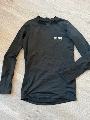 Svart långärmad Select Procare tröja 10/12år - Svart långärmad tröja från Select Procare med diskret logga på bröstet. Tröjan är tillverkad i ett stretchigt syntetmaterial som passar perfekt för träning eller sport. Den har en enkel design med rund hals och smal passform.