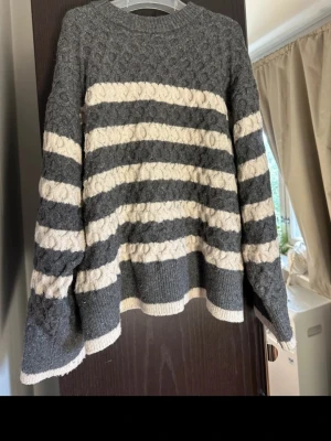 Gråvit stickad randig tröja - Mysig stickad tröja i grått och vitt med breda horisontella ränder. Tröjan har rund hals och långärmade ärmar, samt ett snyggt kabelstickat mönster som ger extra struktur. Perfekt för kyliga dagar och enkel att matcha med jeans eller kjol.