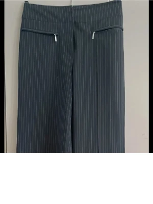 Randiga kostymbyxor från KappAhl - Snygga mörkgrå kostymbyxor från KappAhl med tunna vita ränder. Byxorna har hög midja, raka vida ben och två dragkedjefickor fram. Materialet är en mix av polyester, viskos och elastan för skön stretch och passform.