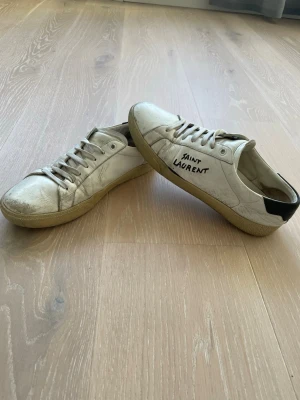 Saint Laurent vita sneakers i skinn - Säljer ett par vita sneakers från Saint Laurent med handskriven logga på sidan. Skorna har snörning, rund tå och beige gummisula. Ovandelen är i skinn och hälen har en svart detalj. Perfekt för dig som gillar klassisk streetstil med designer-touch.