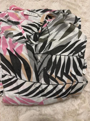 Blommig långklänning från Masai XXL - Säljer en långklänning från Masai i storlek XXL med ett färgstarkt blommigt mönster i svart, rosa, beige och vitt. Klänningen har v-ringning och är tillverkad i ett lätt och luftigt material, perfekt för varma dagar.