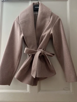 Rosa omlott kappa från ONLY - Säljer en rosa kappa från ONLY i omlottmodell med bred krage och knytbälte i midjan. Jackan har lång ärm och är tillverkad i ett mjukt ullblandat material som ger en lyxig känsla. Perfekt för dig som vill ha en clean och stilren look.