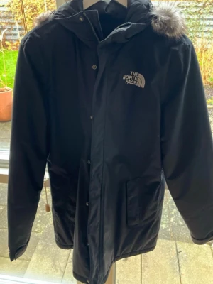 Svart parkajacka från The North Face - Parkajacka i svart med huva och snygg fuskpälsdetalj. Jackan har dragkedja och knappar framtill, två stora fickor och logga på bröstet. Perfekt för kalla dagar och har en clean, stilren look som funkar till många outfits.