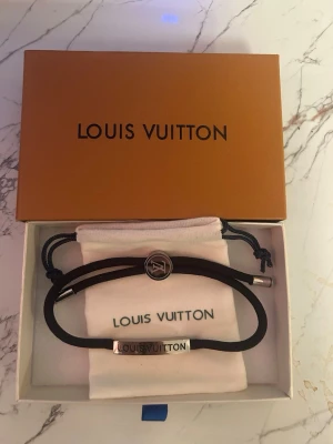 Louis Vuitton armband - Stilrent svart armband från Louis Vuitton med silverfärgade detaljer och logotyp på metallbrickan. Armbandet har justerbar passform med rund låsdetalj och levereras i originalpåse och ask. Perfekt accessoar för dig som gillar lyxiga och minimalistiska smycken.