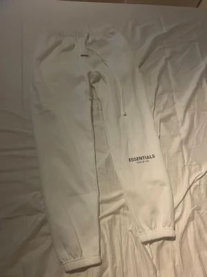 Essentials byxor - Säljer ett par vita mjukisbyxor från Essentials Fear of God med tryck på benet och lapp vid midjan. Byxorna har elastisk midja med snörning och muddar vid bensluten. Perfekta för chill och streetwear. Materialet är mjukt och skönt.