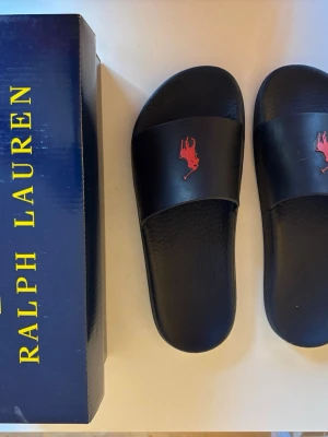 Marinblå  slipin sandaler från Polo Ralph Lauren storlek 40 - Marinblå slipin sandaler från Polo Ralph Lauren med röd logotyp på ovansidan. Storlek 40. Kommer med originalkartong. Inköpspris 895 kr. Jättefint skick!