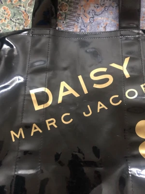 Svart shoppingväska Marc Jacobs Daisy - Cool svart shoppingväska från Marc Jacobs Daisy med glansig finish och stora guldiga blommor på framsidan. Väskan har två rejäla handtag och tryckt logga i guld. Materialet är syntet (PVC) och den är rymlig för allt du behöver ha med dig. Det finns mindre defekter , men inget som direkt stör💖🌸