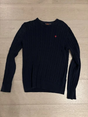 Mörkblå stickad tröja från Morris - Säljer en mörkblå stickad tröja från Morris med kabelstickat mönster och rund hals. Tröjan har ett litet rött broderat emblem på bröstet och långa ärmar. Perfekt för dig som gillar klassisk och stilren design.