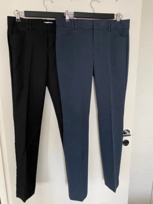 Kostymbyxor Calvin Klein, blå & svarta - Två par stilrena kostymbyxor från Calvin Klein i storlek 6. Ena paret är mörkblå och det andra är svart. Byxorna har raka ben, bälteshällor och är tillverkade av polyester, rayon och spandex för en skön och smidig passform. Perfekta för en clean och snygg look.