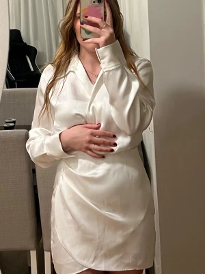 Vit omlottklänning från Zara - Säljer en stilren vit omlottklänning från Zara i storlek XS. Klänningen har långärmade skjortärmar, krage och en snygg omlottdetalj framtill. Materialet är mjukt och har en lätt glansig finish som ger en lyxig känsla. Perfekt för dig som gillar minimalistisk och chic stil.